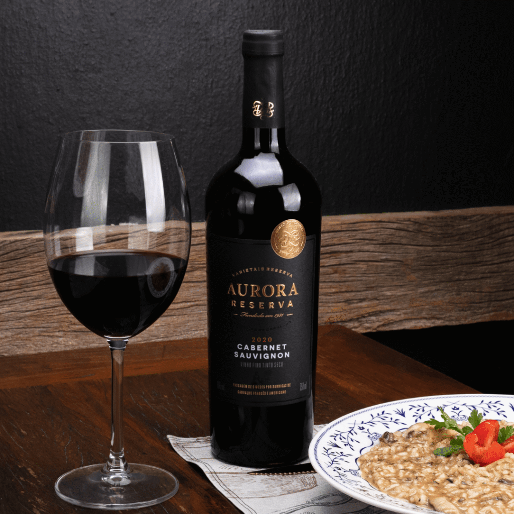 Taça de vinho tinto servida ao lado de uma garrafa Aurora Reserva Cabernet Sauvignon 2020 e um prato de risoto