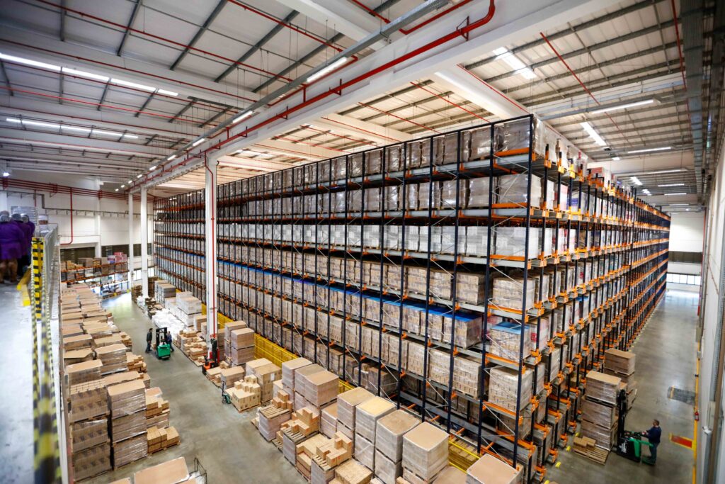 Interior de um grande centro de distribuição com prateleiras altas cheias de caixas e pallets organizados.