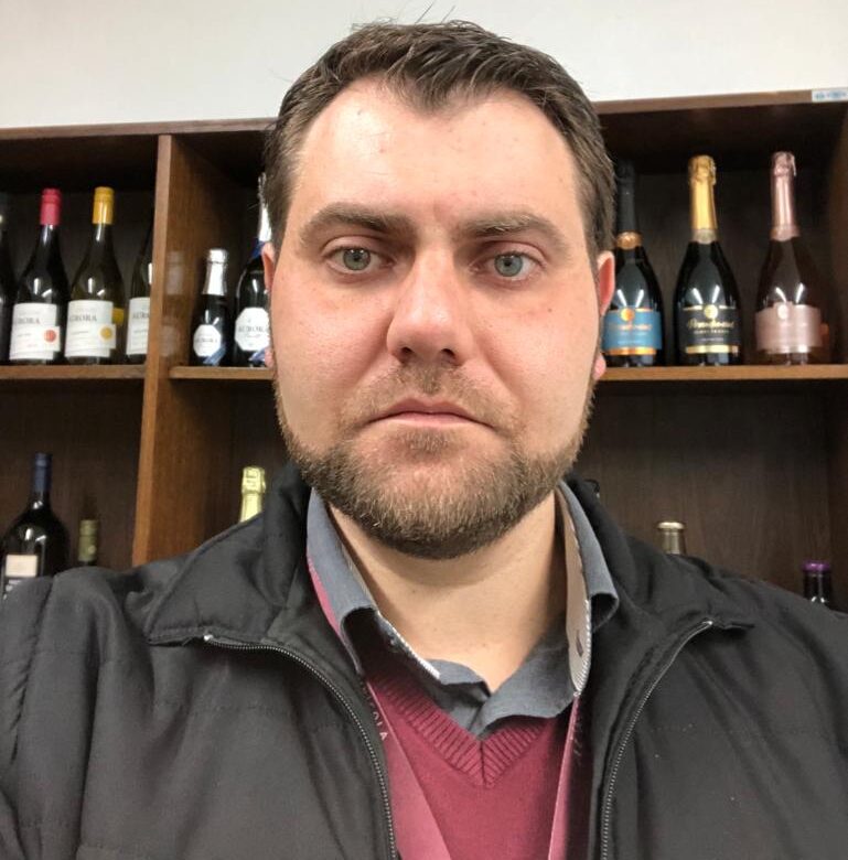 Homem de barba curta e olhos claros olha para a câmera, em frente a uma estante com garrafas de vinho e espumante.