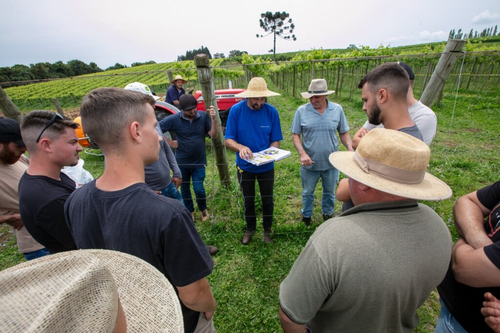 Grupo de agricultores reunidos em campo de cultivo, ouvindo um técnico que apresenta informações em uma prancheta.