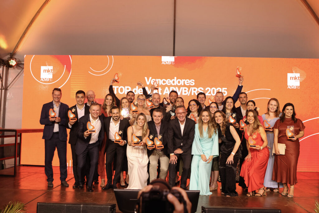 Um grande grupo de pessoas posa sorridente em um palco, exibindo troféus de premiação.