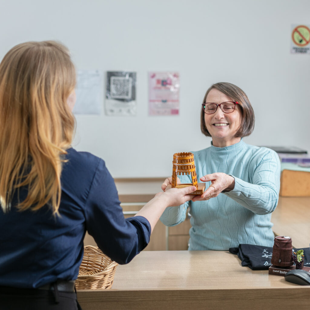 Mulher idosa sorridente entrega um objeto artesanal de madeira a outra pessoa em um balcão.