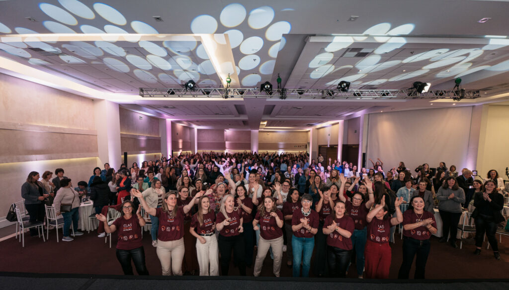 Grande auditório lotado com dezenas de mulheres em pé, batendo palmas e celebrando.