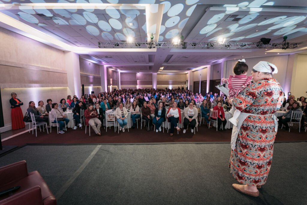Grande auditório lotado com dezenas de mulheres em pé, batendo palmas e celebrando.