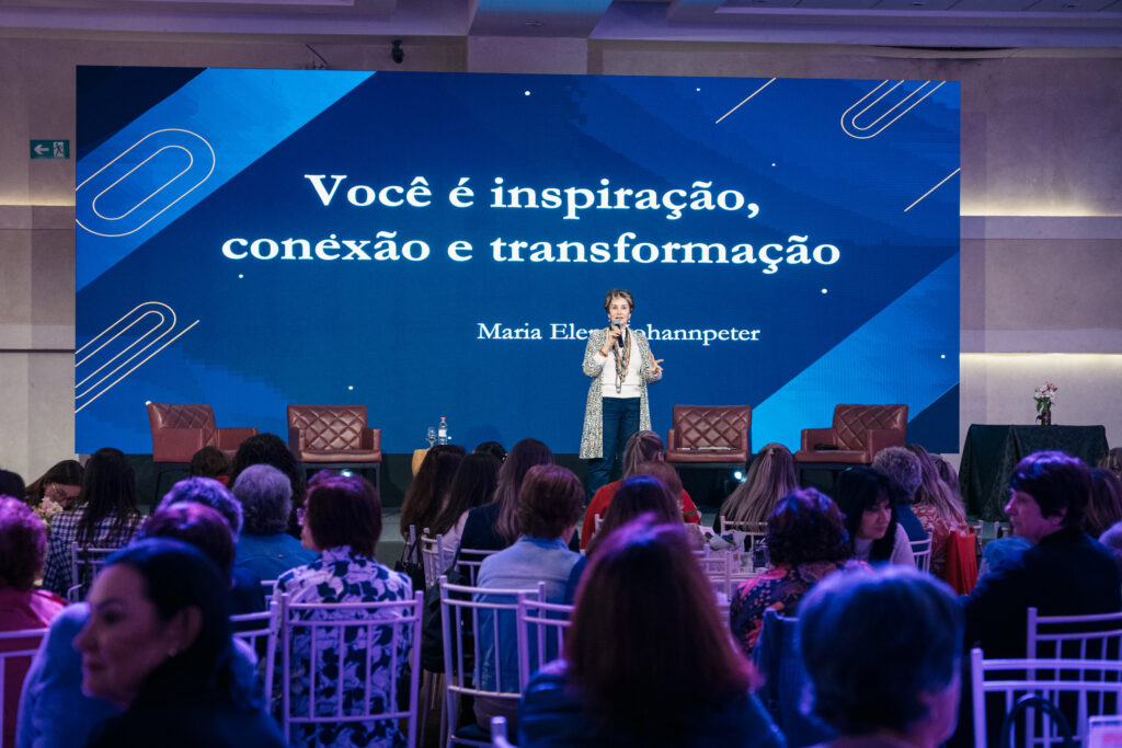 Uma mulher está em pé no palco, falando ao microfone para uma plateia.