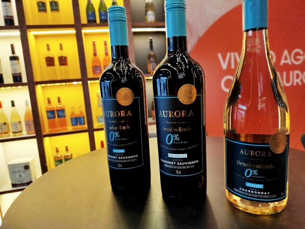 Três garrafas de vinho desalcoholizado Aurora sobre uma mesa, com prateleiras de bebidas iluminadas ao fundo.
