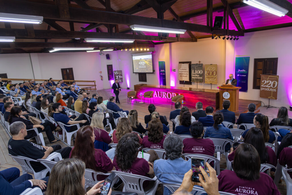 Palestra corporativa na Vinícola Aurora com auditório cheio, palco iluminado e banners institucionais destacando marcos da cooperativa.
