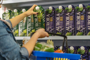 Pessoa pegando uma embalagem de suco de uva Aurora em uma prateleira de supermercado, com várias unidades do produto ao fundo e uma cesta de compras à frente.