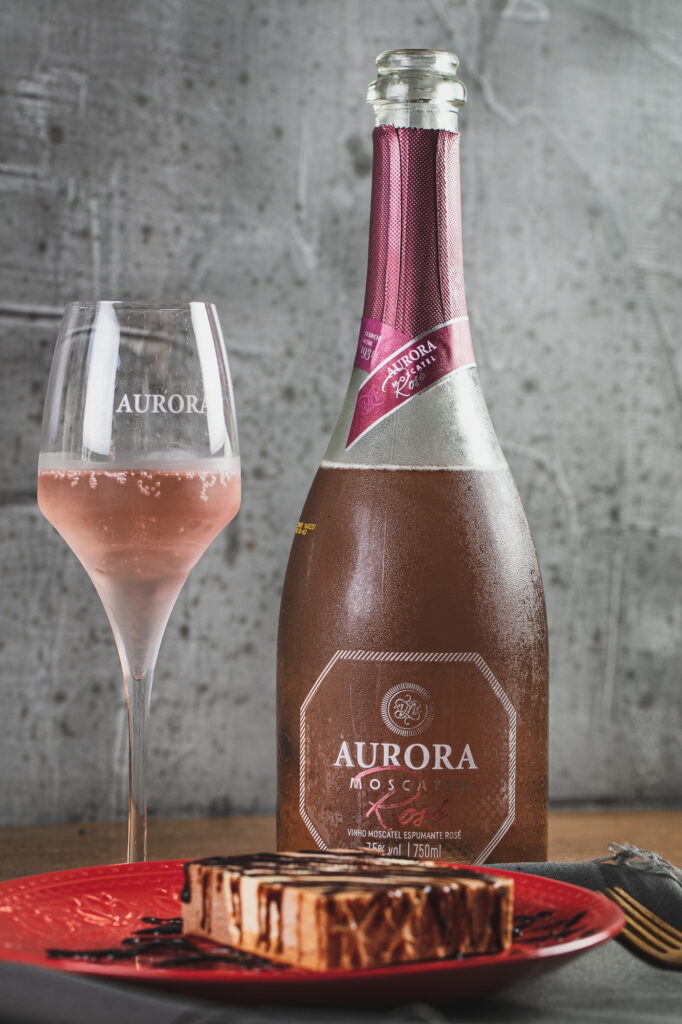 Garrafa e taça de Aurora Moscatel Rosé ao lado de sobremesa com calda de chocolate, em composição elegante sobre fundo cinza.