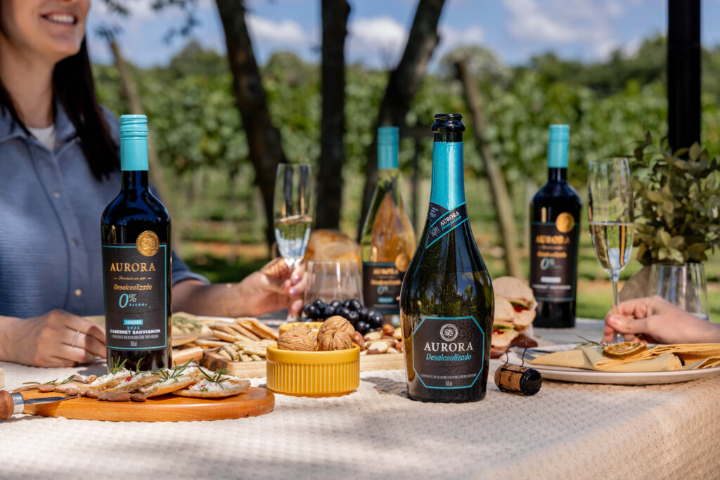 Mesa ao ar livre com vinhos Aurora desalcoolizados, petiscos e pessoas brindando em vinhedo.