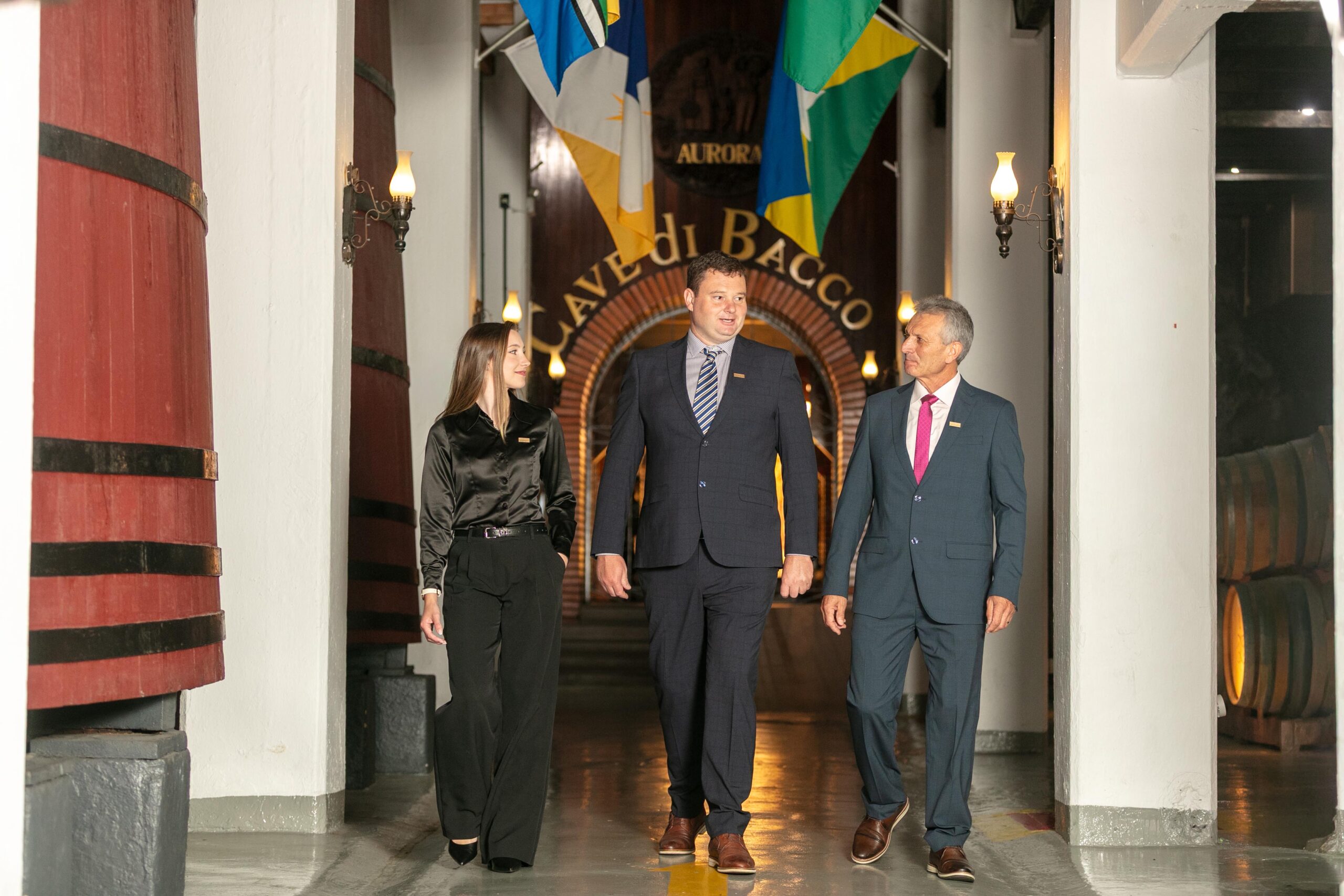 Três pessoas em trajes formais caminham em corredor de vinícola.