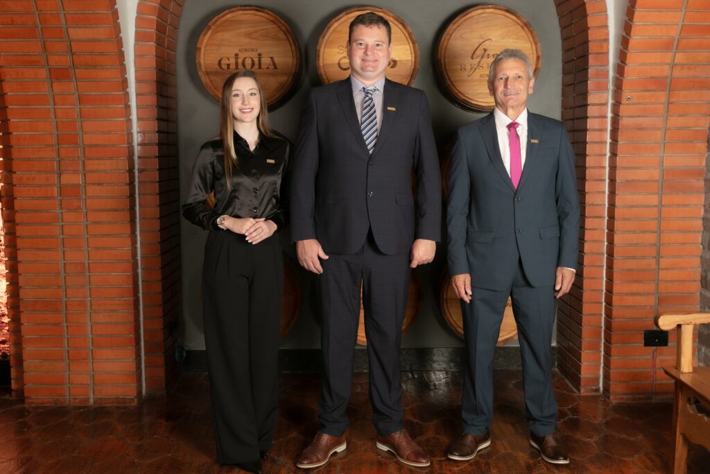 Três profissionais posam em vinícola diante de barris de vinho.