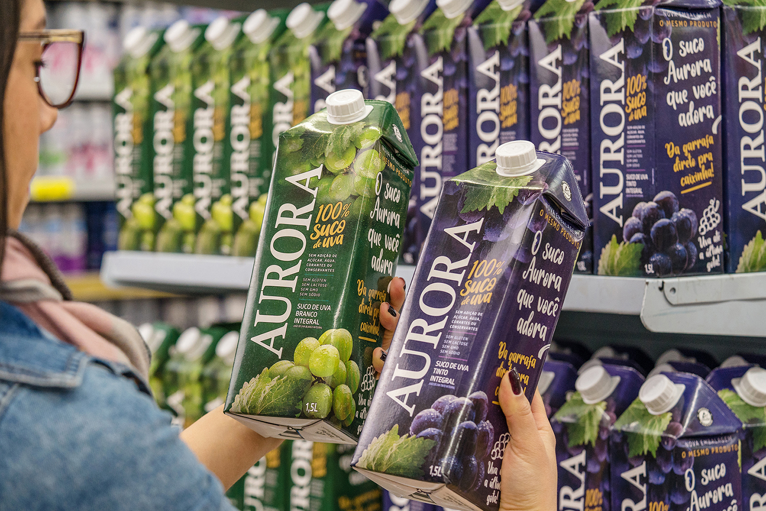 Pessoa comparando embalagens de suco de uva Aurora em supermercado.
