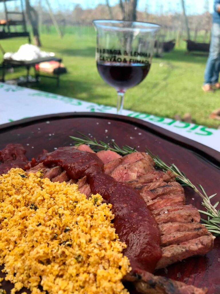 Prato com carne grelhada, farofa e taça de vinho ao fundo.