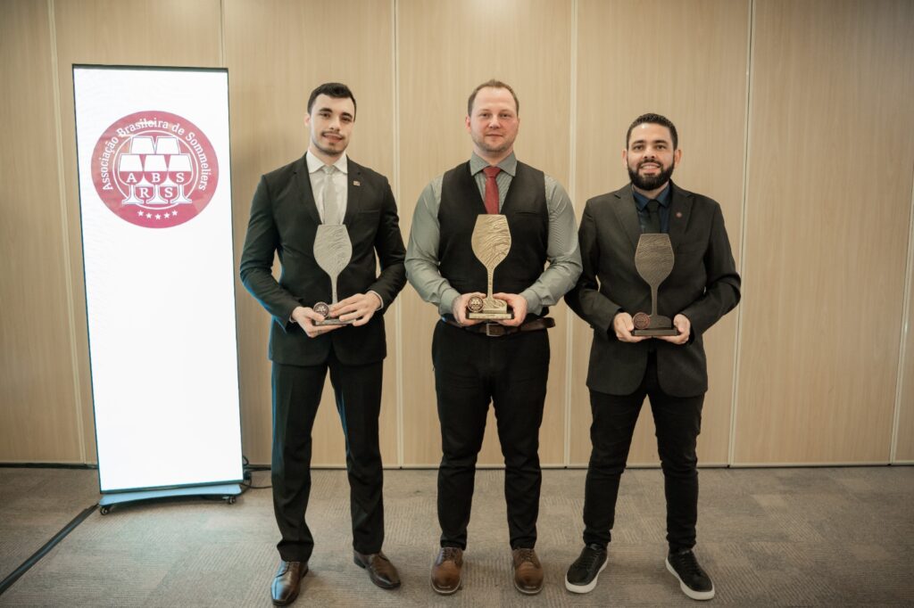 A imagem mostra três homens posando lado a lado em um ambiente formal, segurando troféus em formato de taça de vinho, em uma cerimônia ligada ao universo da sommellerie. Todos estão vestidos de maneira elegante, com trajes sociais em tons escuros, transmitindo profissionalismo e sofisticação.

Ao lado esquerdo da foto há um painel vertical com o logotipo da Associação Brasileira de Sommeliers do Rio Grande do Sul (ABS-RS), indicando que se trata de uma premiação ou reconhecimento promovido pela entidade.

O homem ao centro segura um troféu maior, sugerindo a primeira colocação, enquanto os outros dois participantes, posicionados à esquerda e à direita, também exibem suas premiações, compondo um pódio simbólico dos destaques do concurso.

O ambiente possui decoração neutra e corporativa, com paredes em madeira clara e iluminação suave. A imagem transmite celebração, reconhecimento profissional e valorização da excelência no universo dos vinhos e da sommellerie.