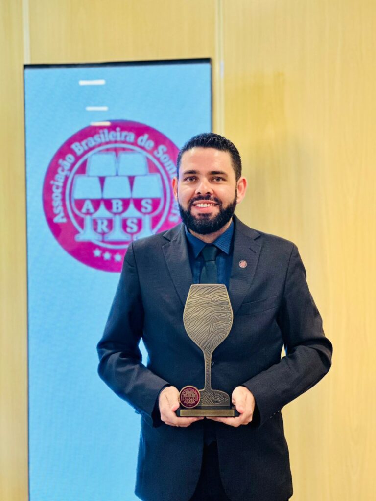 A imagem mostra um homem sorridente, vestido com traje social escuro, camisa preta e gravata, segurando um troféu em formato de taça de vinho. Ele aparece posicionado em frente a um painel da Associação Brasileira de Sommeliers do Rio Grande do Sul (ABS-RS), indicando um contexto ligado ao universo da sommellerie e premiações do setor.

O ambiente é elegante e corporativo, com fundo em tons claros de madeira, transmitindo formalidade e sofisticação. A expressão do homem demonstra orgulho e satisfação pela conquista. O troféu reforça a ideia de reconhecimento profissional e destaque na área dos vinhos.

A composição da imagem transmite credibilidade, excelência profissional e celebração de uma conquista importante no segmento da sommellerie.
