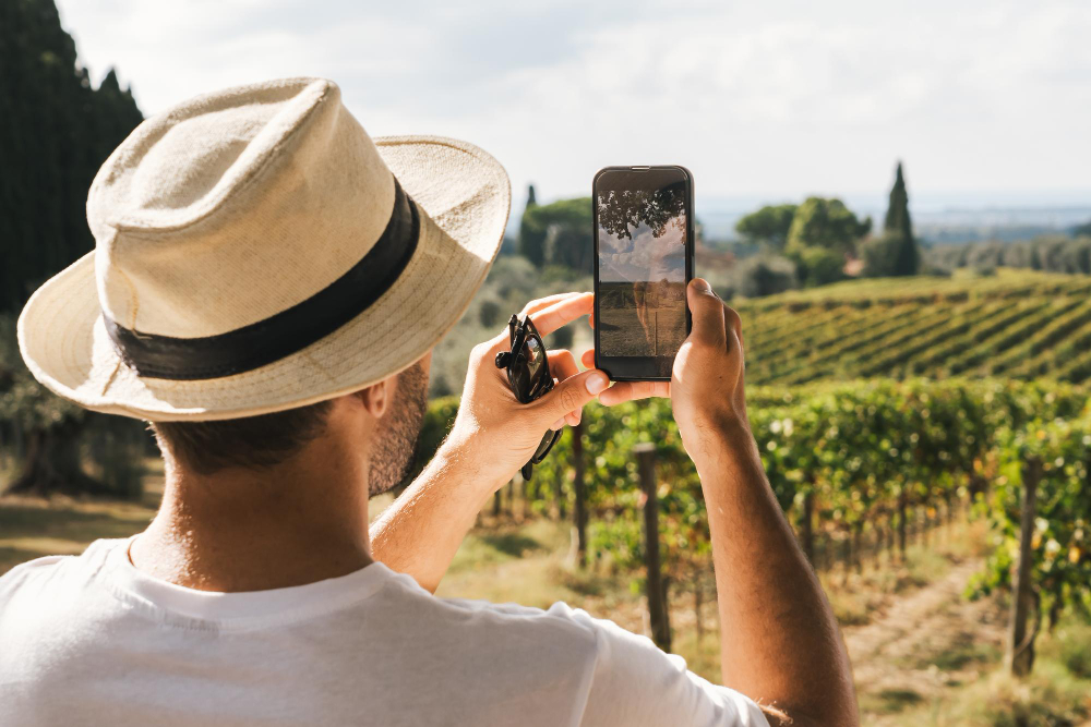 Pessoa usando chapéu fotografando um vinhedo com smartphone durante visita a uma região produtora de vinhos.