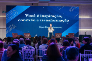 Uma mulher está em pé no palco, falando ao microfone para uma plateia.