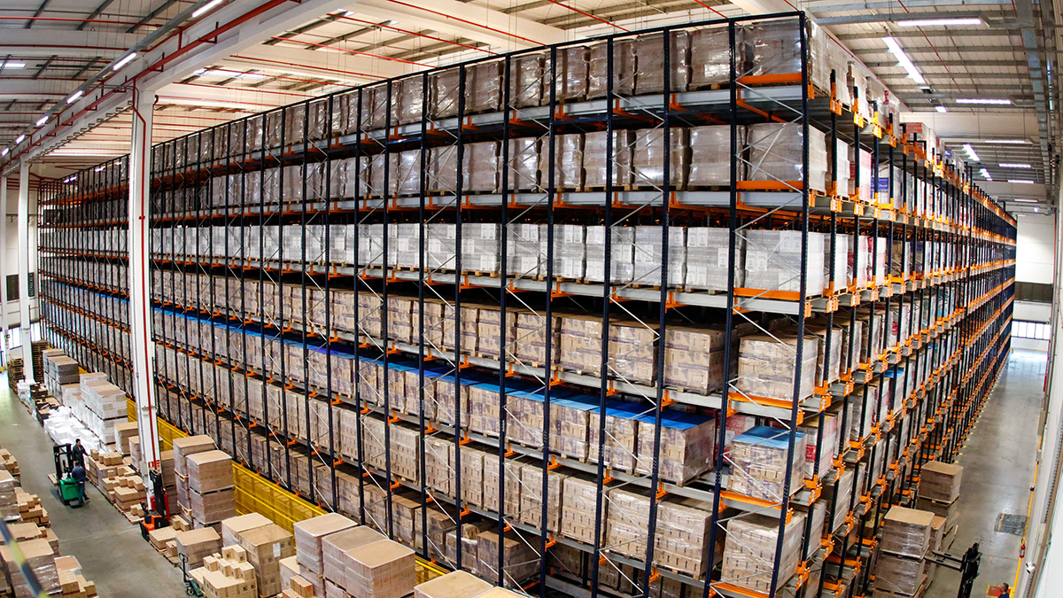 Armazém industrial amplo com estantes altas e centenas de pallets empilhados, indicando grande capacidade de estocagem e operação logística. No chão, caixas e pallets organizados e empilhadeiras em movimento.